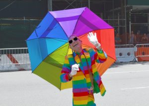 man op straat in regenboogpak met witte handschoenen, zonnebril en regenboogparaplu